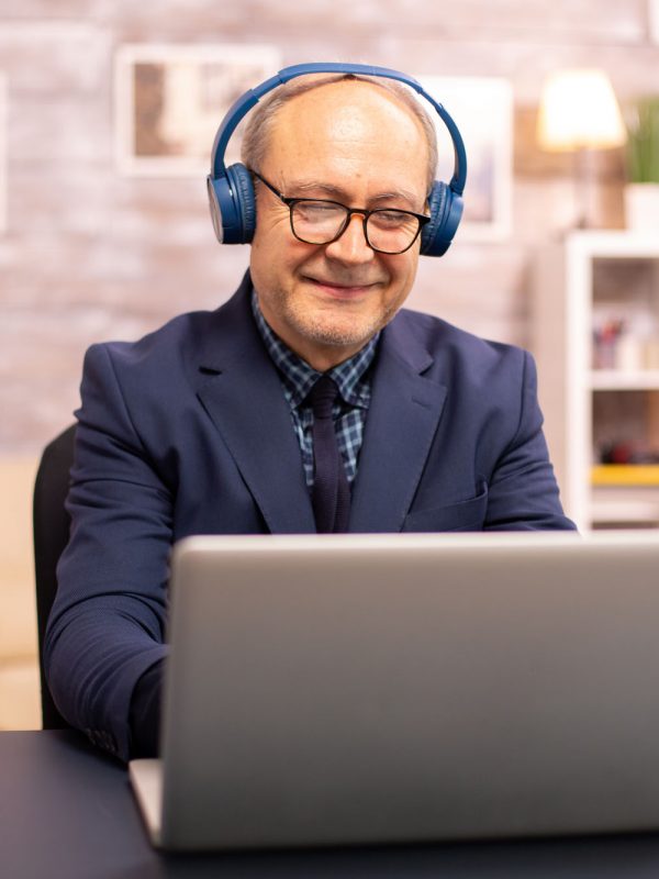 Elderly man in his 60s with headphones on his head listening to music and working on a modern laptop